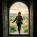 Woman walking through stone archway into green fields with mountains and sunset sky