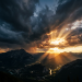 Sunset rays piercing through dark storm clouds over a mountainous valley with a river